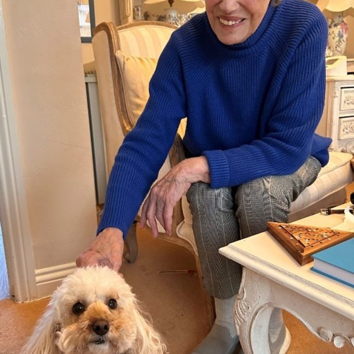 An older lady sits on a chair petting a small white dog